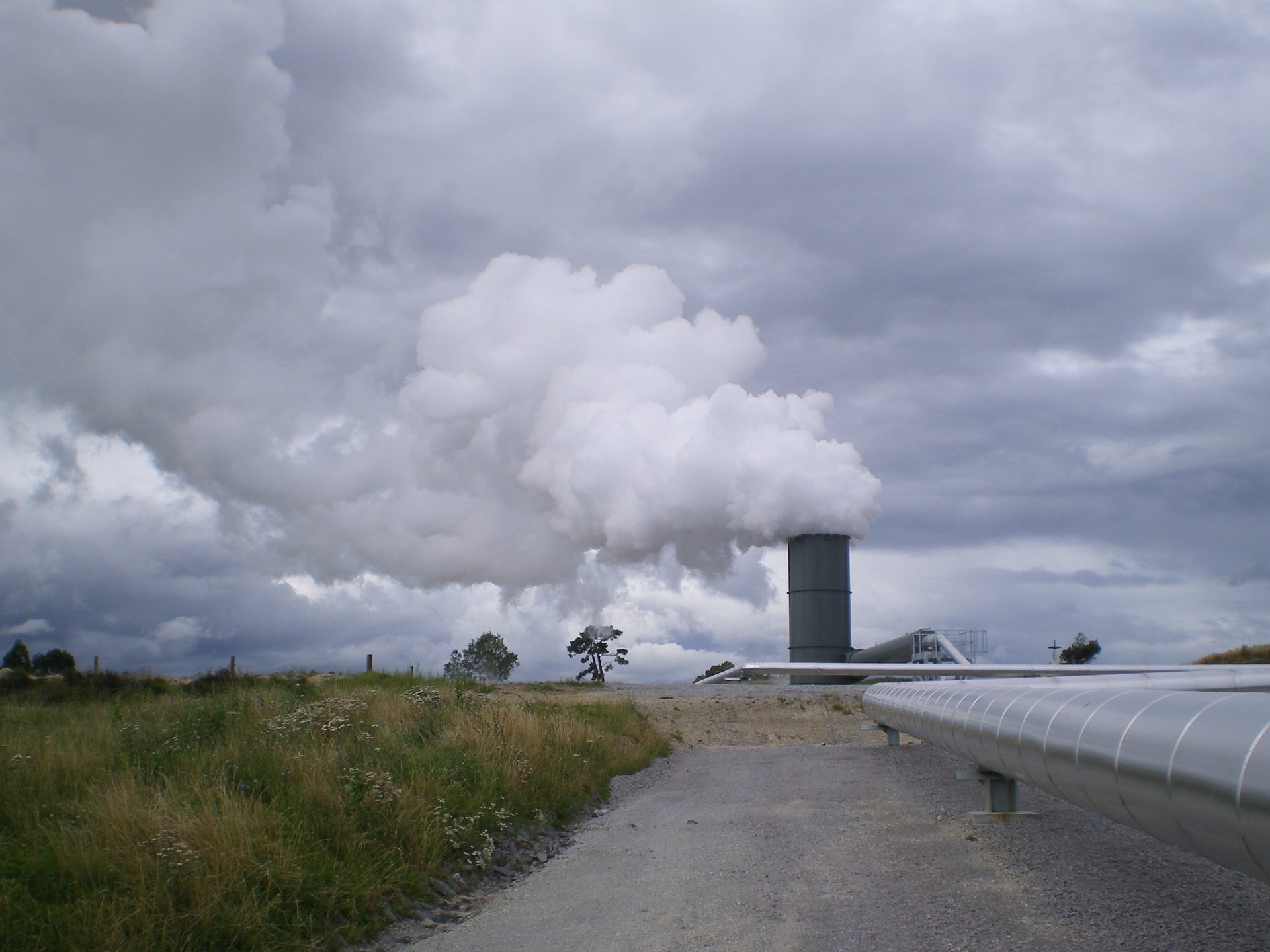 White steam venting from a geothermal stack into heavy cloud beside an insulated pipeline