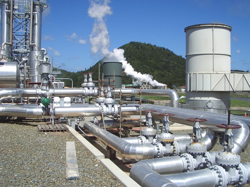 NTGA clean steam plant at Kawerau: insulated geothermal pipework and valves, a steam plume and a bush-covered hill