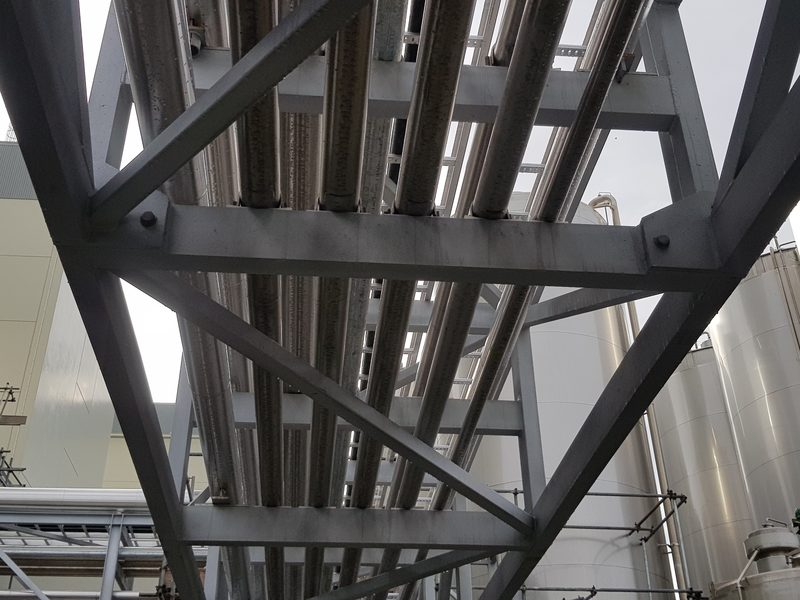 Looking up into a bolted, cross-braced steel pipe bridge carrying process pipework, with silos behind